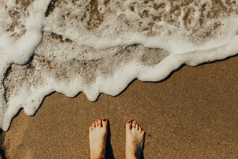an overhead shot of a persons feet in wet coarse sand next to small foaming waves, feet in the wet sand 4k