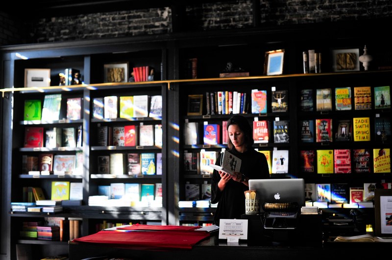 a woman skimming through a book while behind the cash register in a bookstore, lost in reading 4k