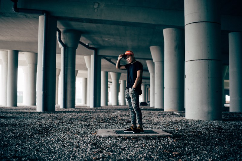 a man holds his hand on the brim of his baseball cap beneath a bridge, hypebeast on a budget 4k