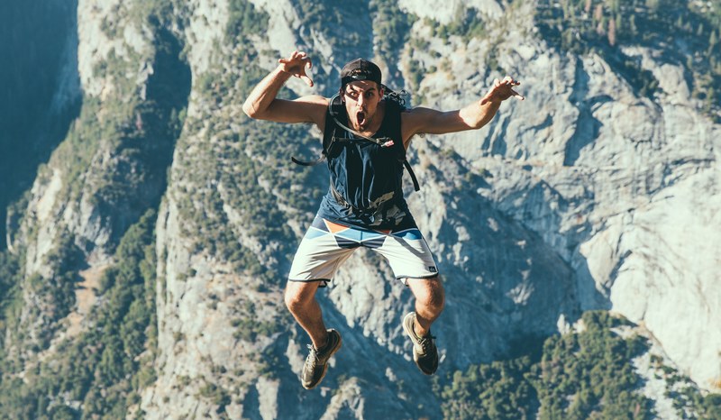 a man jumping in the air wearing shorts a tank top a water backpack and sneakers on a hike in the mountains, man skydiving by cliffs 4k