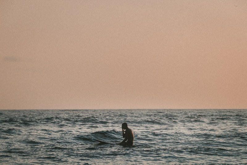 a surfer sitting on their surfboard in the ocean underneath a dark orange sky, surfer 4k