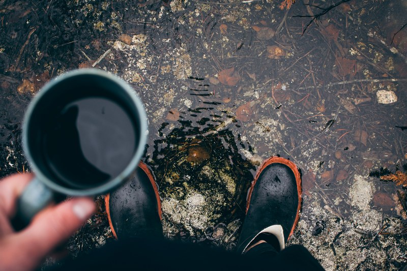 a person wearing rain boots stands in a puddle looking down and holding coffee, morning coffee 4k