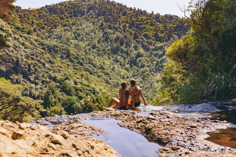 two girls in swimsuits sitting on the edge of a small rocky waterfall in piha forest, sunbathing in a piha forest 4k