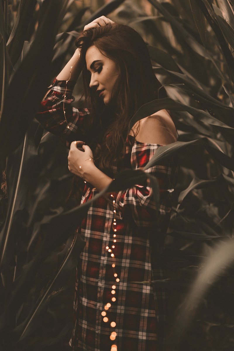 a woman in a plaid off the shoulder dress holds a string of lights among tall plants, woman holding string of lights 4k