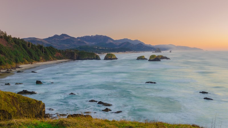 ocean washing on a sandy beach with green cliffs in ecola state park at sunset, shoreline at sunset 4k