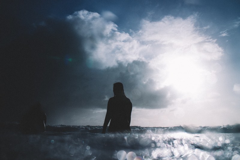 a silhouette shot of two surfers sat on their boards in the calm sea at piha beach as a cloud passes in front of the sun, surfers sitting on boards 4k