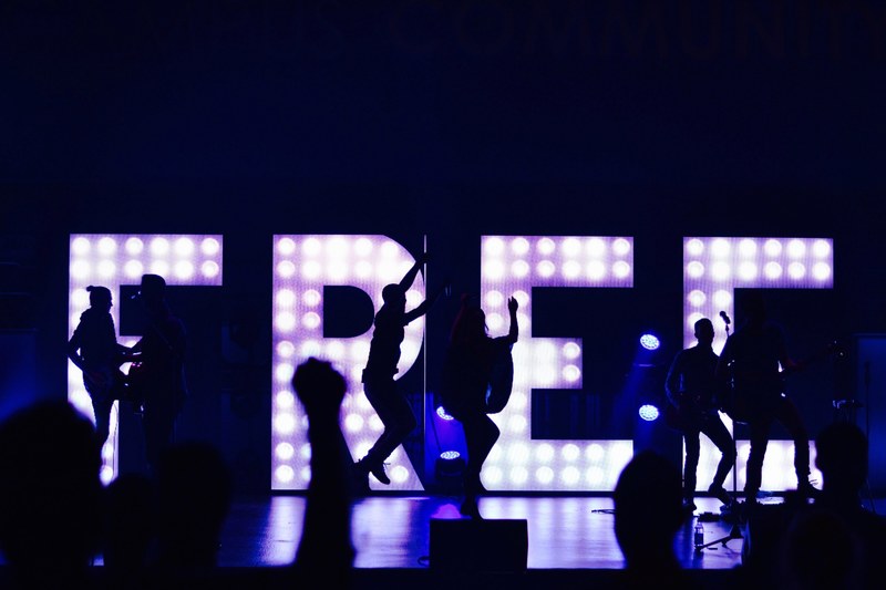 silhouettes of band members performing and jumping up in front of lights arranged to spell out free, concert stage performance and free 4k