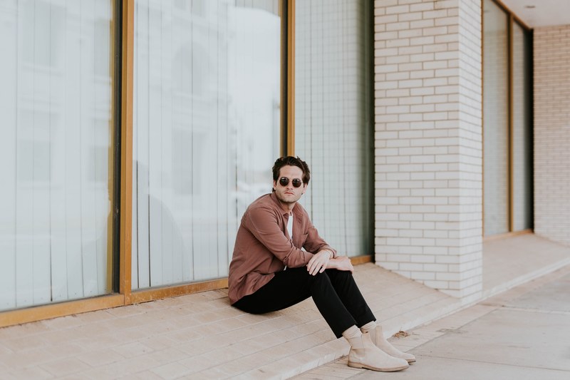 a man in sunglasses sits on a brick ledge next to plate glass windows, man on brick window ledge 4k