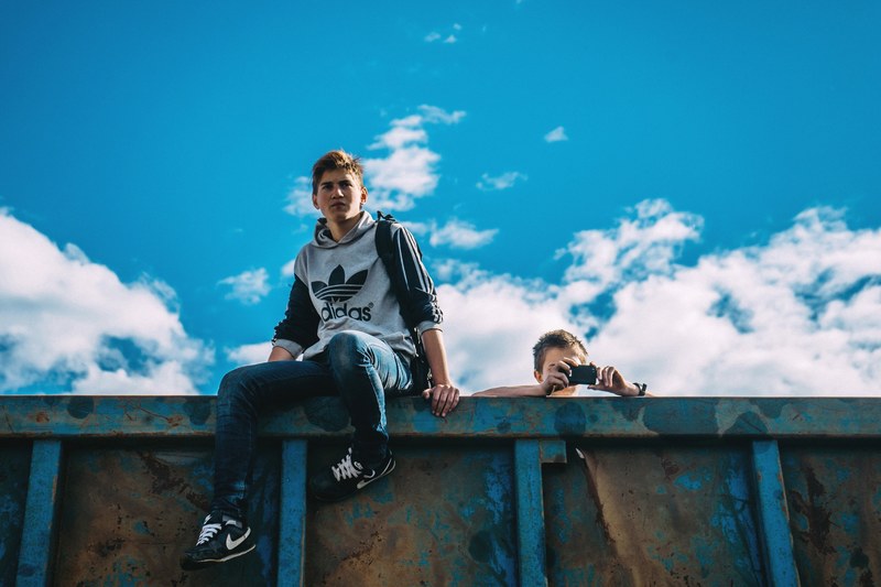 two men on a rusted wall against a blue sky one taking photos with an iphone, men on rusted wall 4k