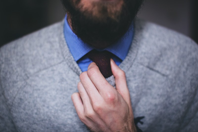 a bearded man in a blue shirt and gray sweater adjusting his tie, man in a sweater adjusting tie 4k