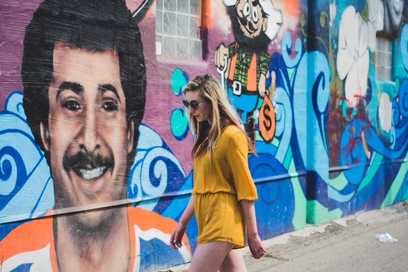 attractive young blonde woman in sunglasses and yellow dress walking past colorful face graffiti artwork, girl in front of mural 4k