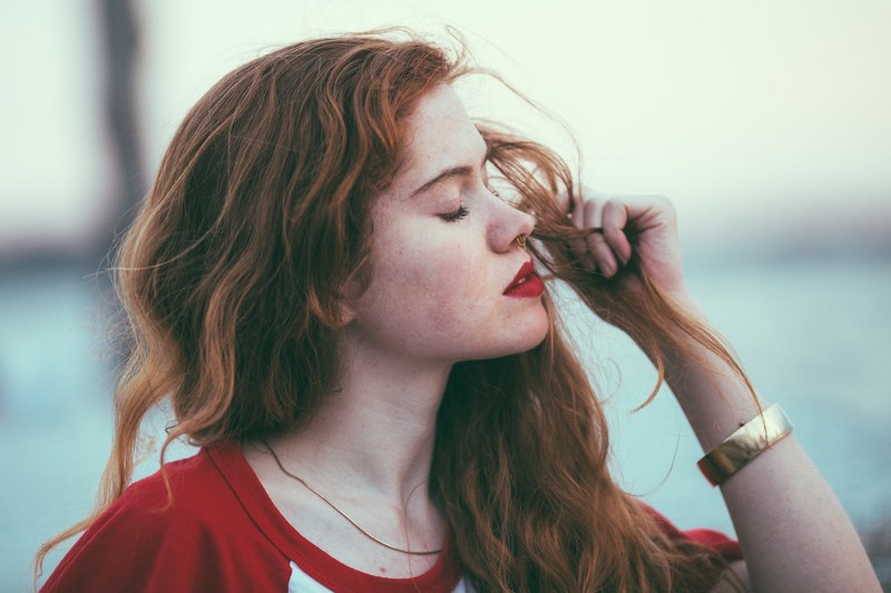 a woman with long red hair and red lipstick holds her hair while closing her eyes, lilian at sunset 4k