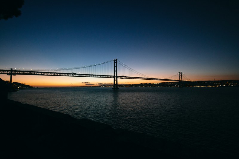 the end of a sunset behind a large suspension bridge over water in lisbon, lisbon suspension bridge sunset_ 4k