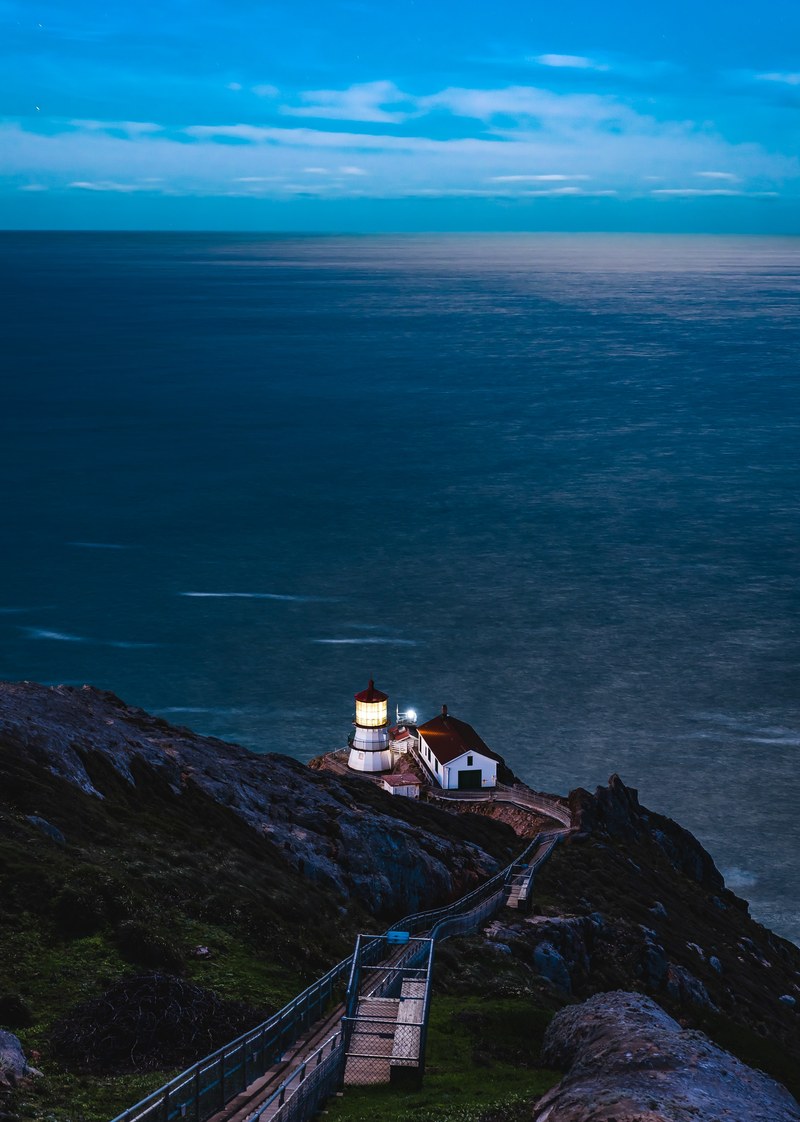 white and red light house with a lit light and barn next to it on a cliff overlooking the sea, night light 4k