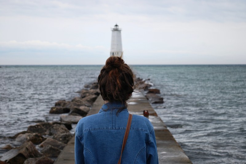 a woman in a bun walks down a rocky pier on the ocean toward a lighthouse, woman walking to lighthouse 4k
