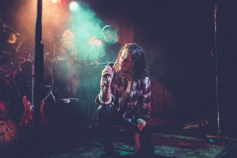 a long haired male singer in a checkered shirt singing into a microphone on one knee, kneeling singer 4k