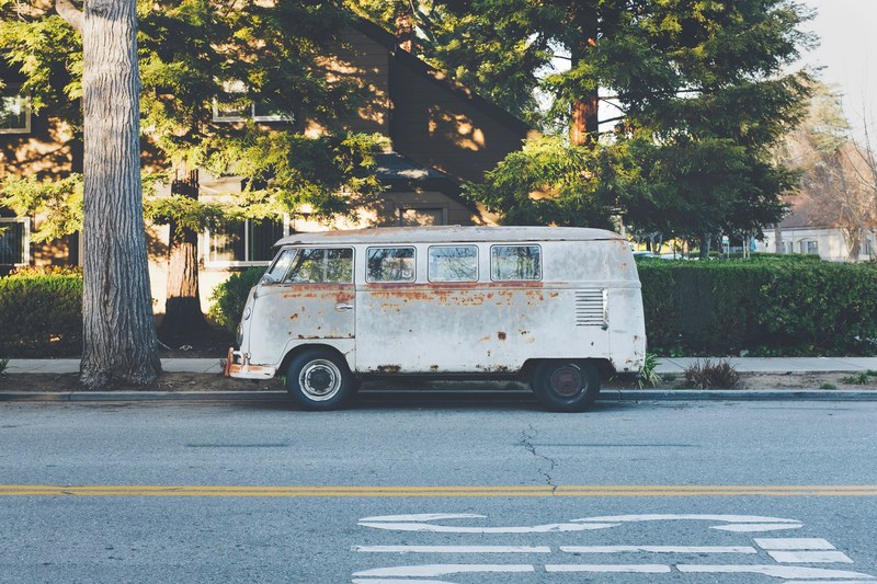 a white rusty vintage volkswagen bus parked on the side of a street by a large tree, hippie bus 4k