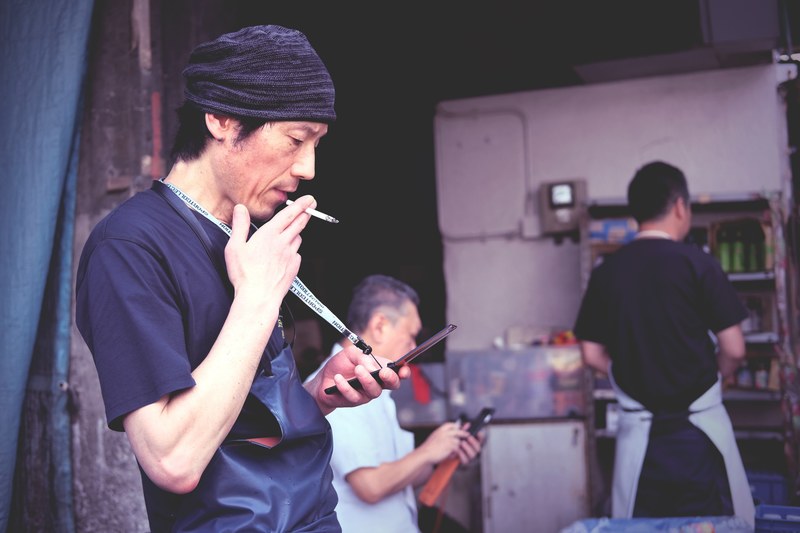 a man is smoking and looking at his phone in the foreground at tsukiji market, _man in apron smokes cigarette 4k