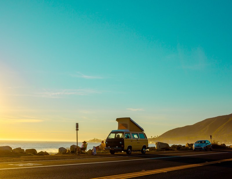 a volkswagen camper van parked at the side of a coastal road with its roof up and two people sitting beside it looking out to sea_ bright clear blue skies at sunset, la to sf 4k