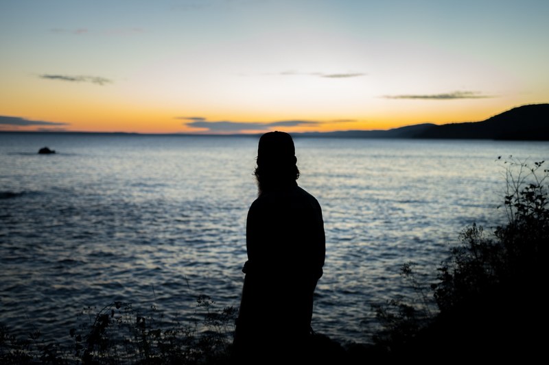 a silhouette of a person looking off into the distance on the shore of a lake at dusk, watching it set 4k