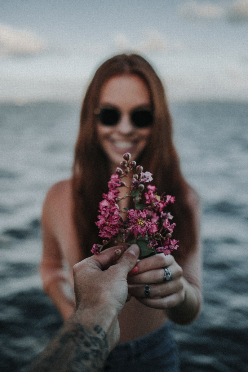 a woman in sunglasses by the water holds out pink flowers to a tattooed mans arm, woman passing flowers to man 4k