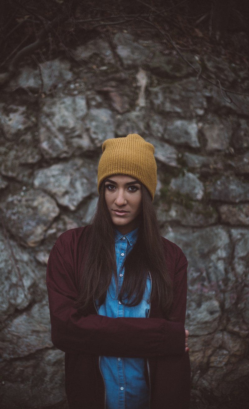 a woman in a brown beanie and maroon jacket against a stone wall in hungary, _look into my eyes 4k
