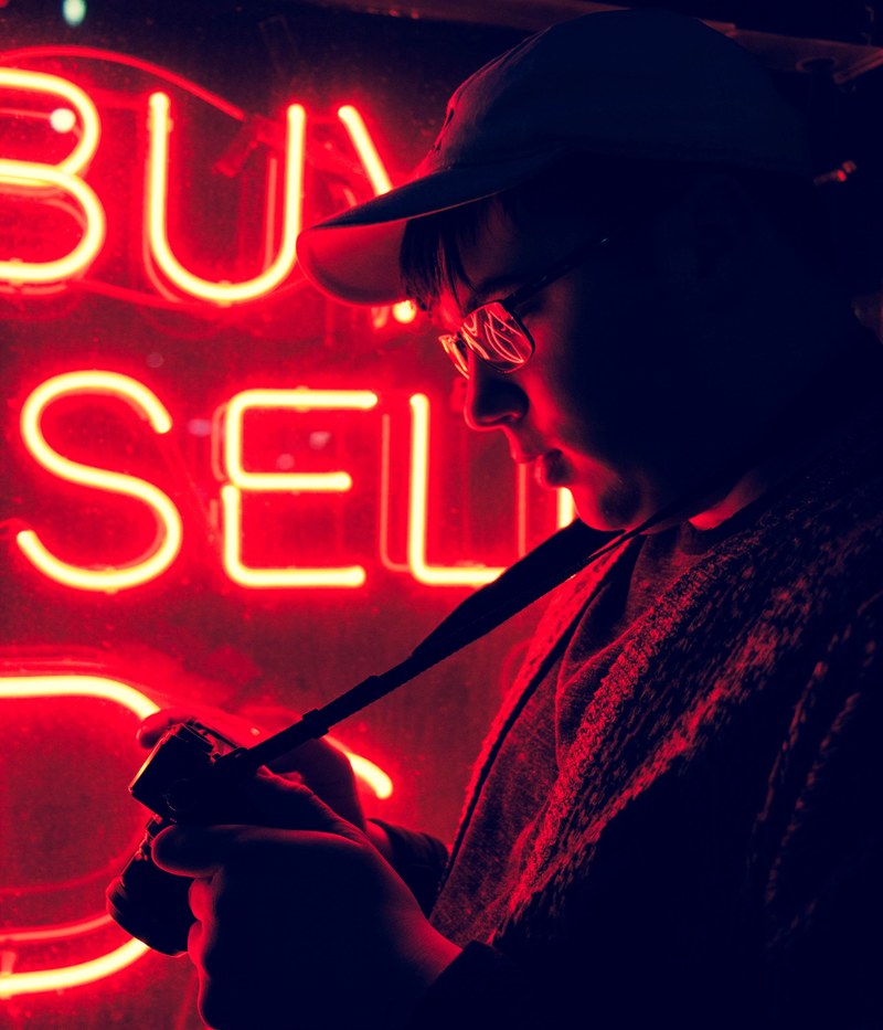 a man in glasses looking at his camera while standing near a red neon, neon_2 4k
