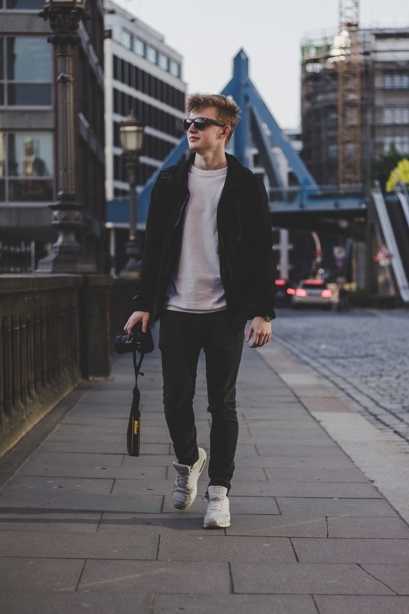 a young man wearing sunglasses walking over a bridge holding a camera in hafencity, man walking with a camera 4k