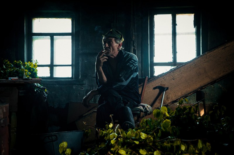 older man sitting by the window in a dimly lit room smoking a cigarette in knyazeva russia, _russian taiga 4k