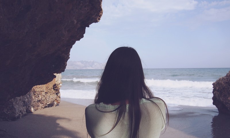 a dark haired woman looking at the crashing sea waves under a cliff, sea breeze under the cliff 4k