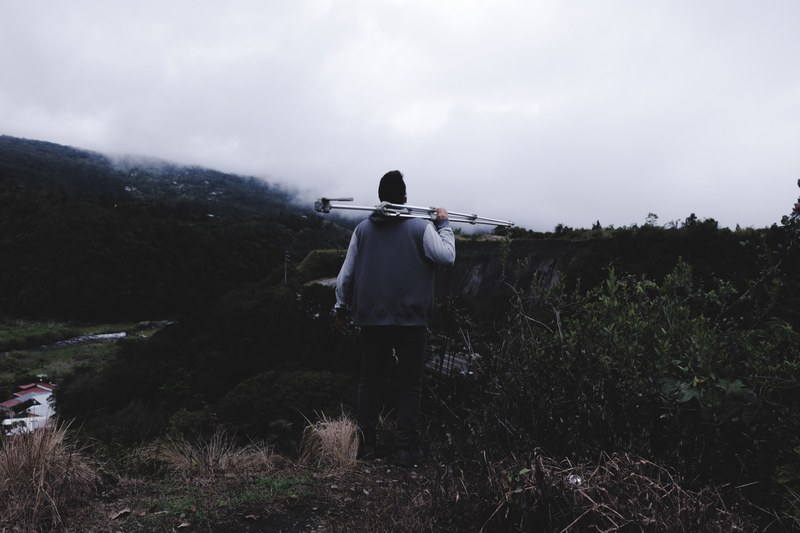 a man with a tripod on his shoulder and a camera in his hand looks off into the wooded landscape, looking for a photo spot 4k