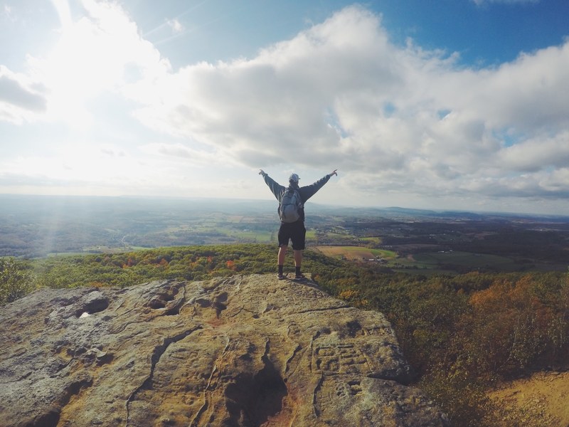 a man with a backpack standing with his hands outstretched on a rocky ledge overlooking green countryside, high above the green plains 4k