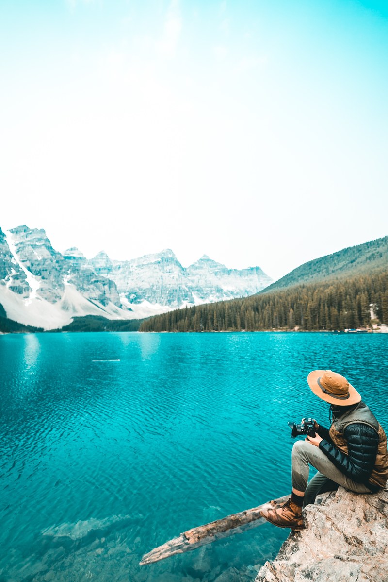 a person sitting on a rock over an azure lake and holding a camera, explore 4k