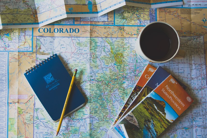 rite in the rain notebook coffee pencil and three atlases sit on a map of colorado, planning the journey 4k