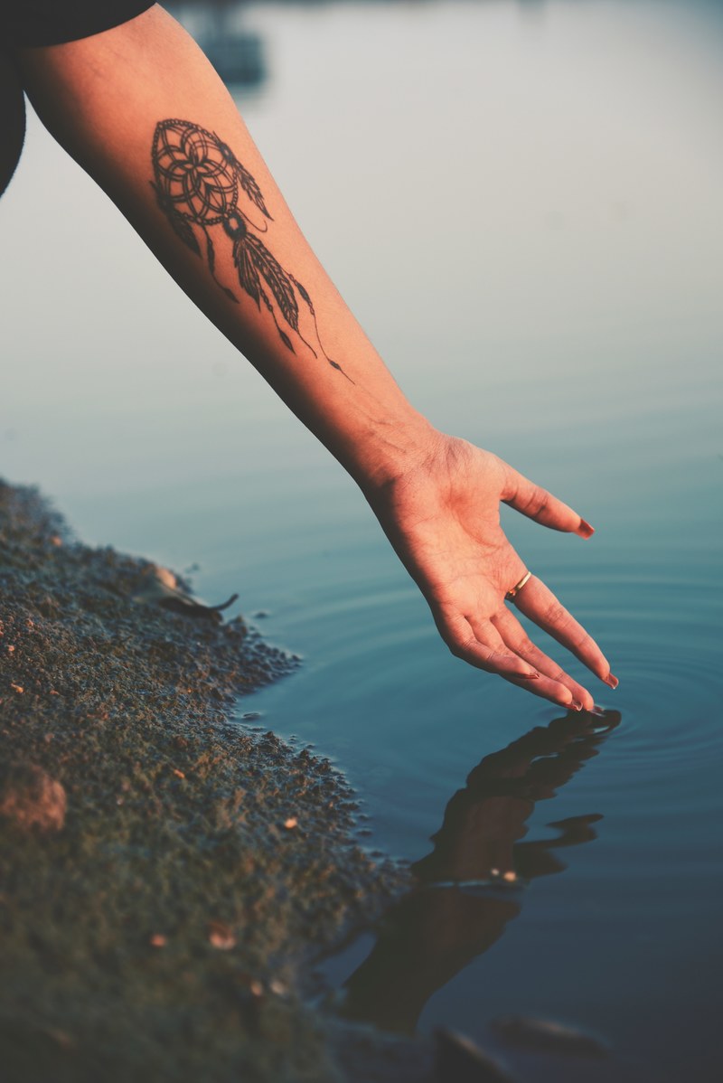 a tattooed woman reaches her arm into calm lake waters, tattooed lake reflections 4k