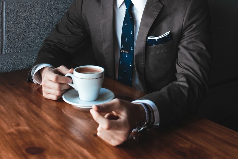 a businessman in a black suit and a tie drinking coffee at a table, damn fine coffee 4k