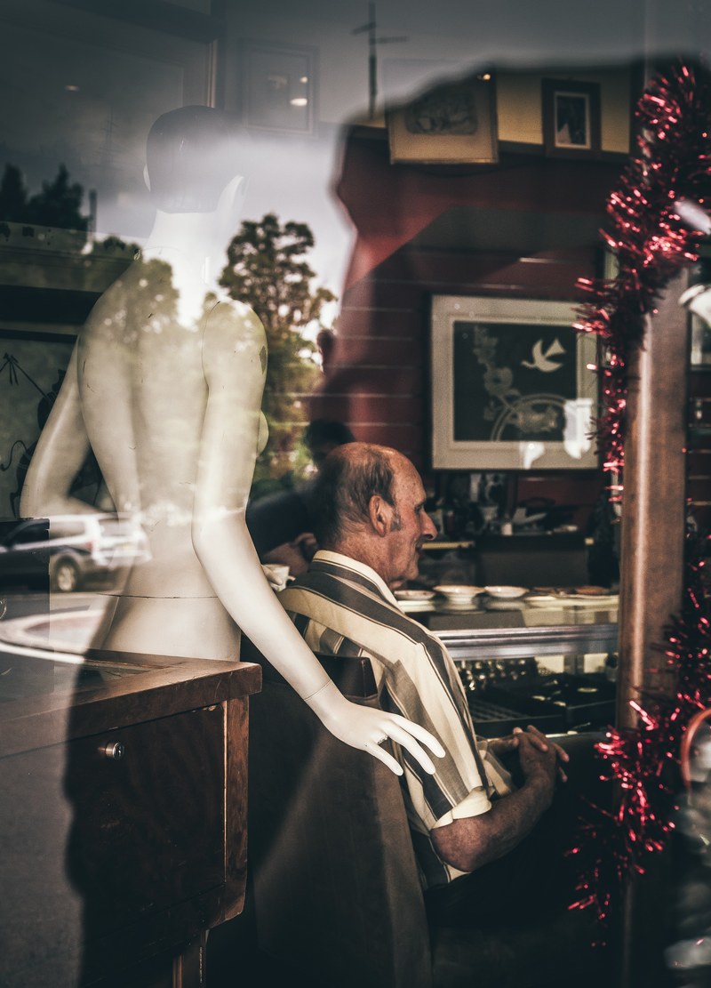 an elderly man sits at a store in katoomba with mannequin behind him the window has reflections of trees, _mannequin behind man 4k