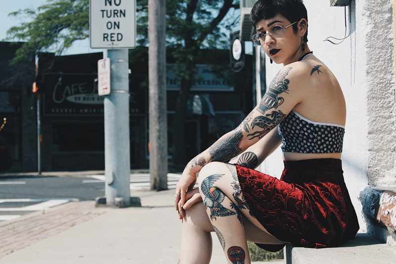 woman with edgy tattoos wearing a skirt sits on a bench outside, showing off her tattoos 4k