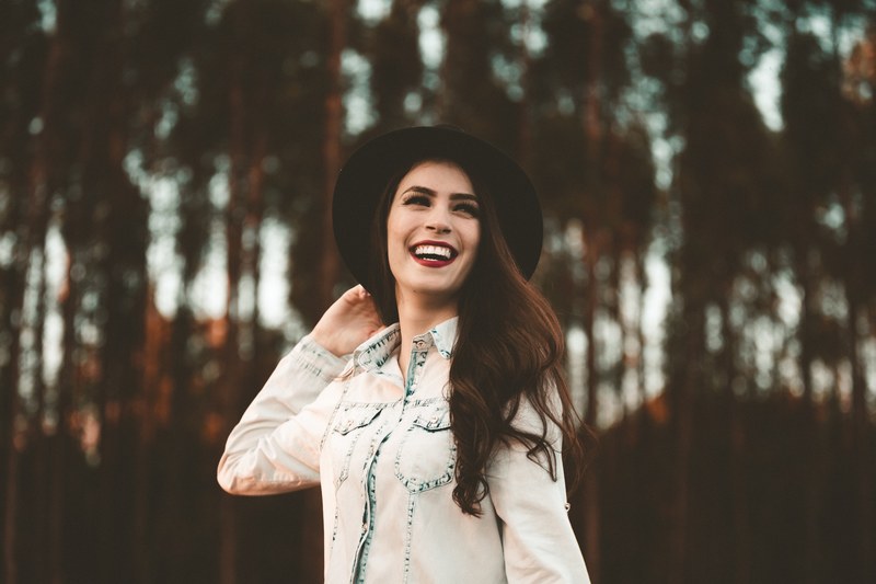 a woman in dark lipstick and a black hat smiles broadly in front of tall trees, woman in hat with tall trees 4k