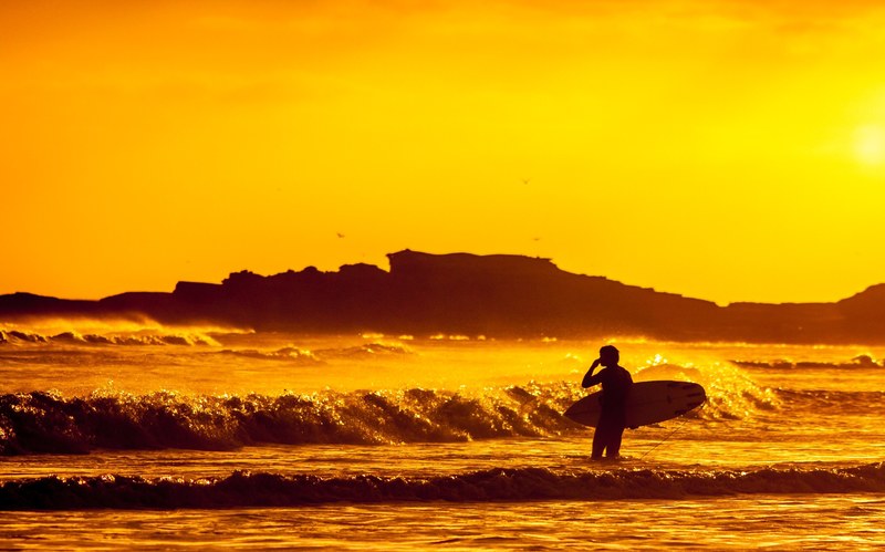 a surfer stood in the sea at sunset holding a board beneath a vibrant orange dusk sky, orange sunset surfer 4k