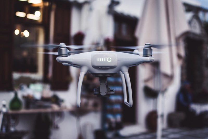 a white drone hovering in front of a white home with brown shutters and doorway beside a patio umbrella, white drone hovering 4k