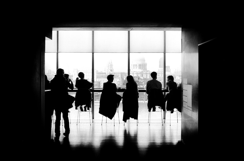 a black and white shot of people sitting on stools at a long table, discussions at a long table 4k