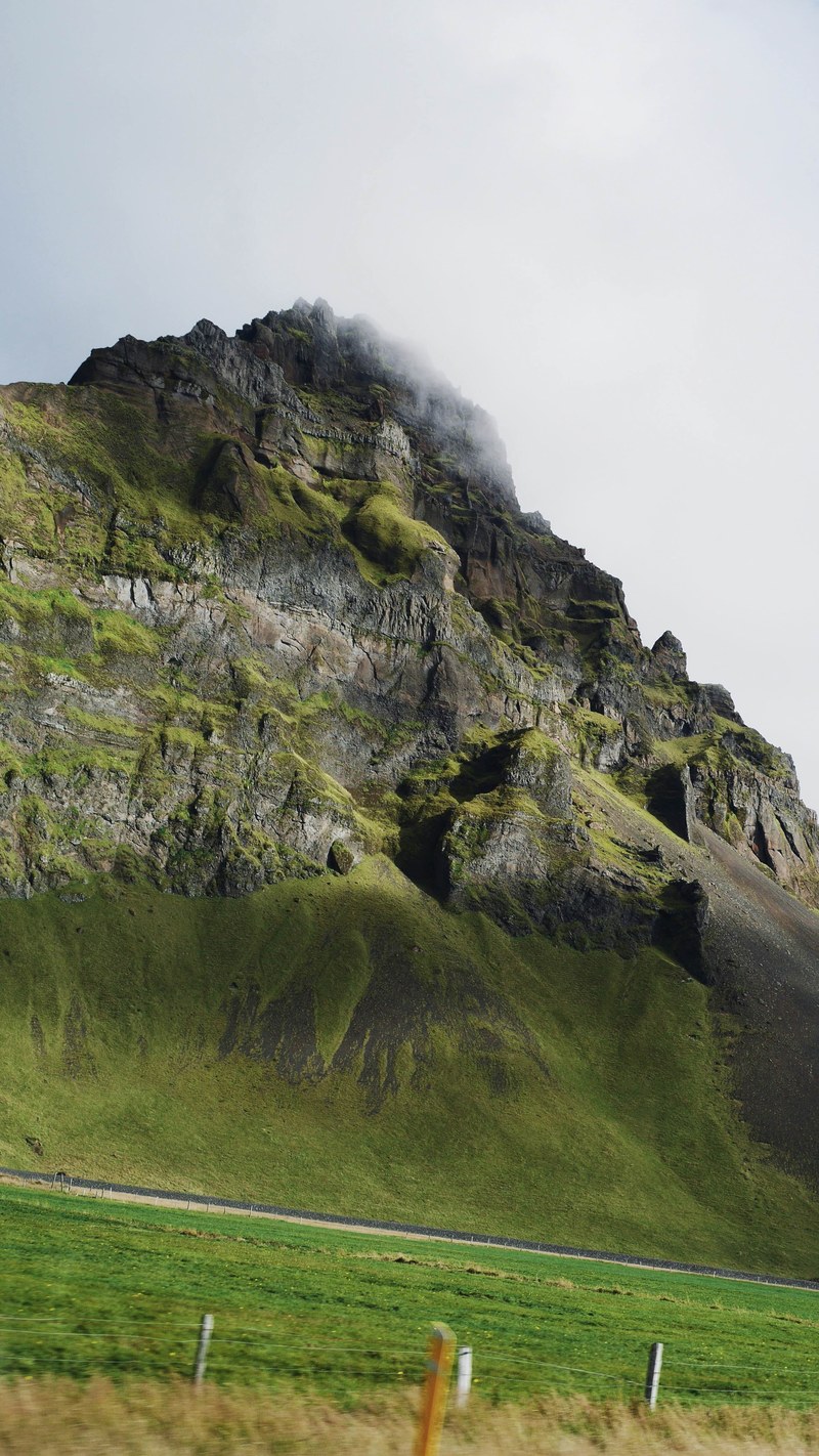 a craggy mountain with its top shrouded in fog rising above an asphalt road, mountain towering over a road 4k