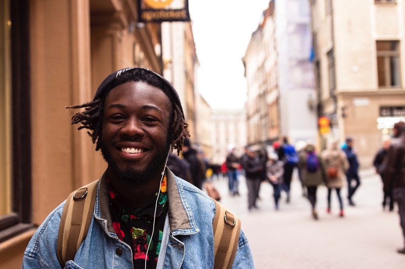 a young man in a jean jacket with a backpack and headphones smiles outdoors in stockholm, stockholm man with headphones 4k