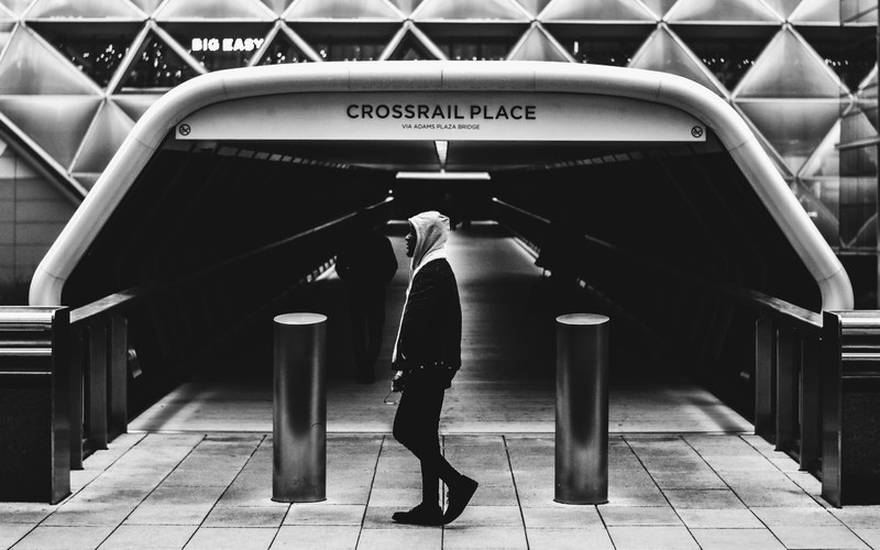 black and white shot of man in grey hood walking near crossrail place tunnel and sign, past the tunnel 4k