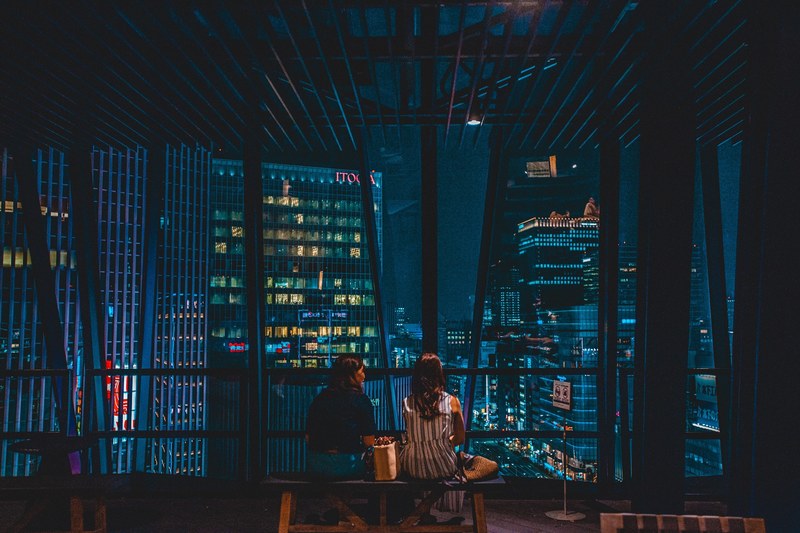 two women sitting on bench near large skyscraper window looking at city skyscrapers at night tokyo, tokyo skyscraper window view 4k