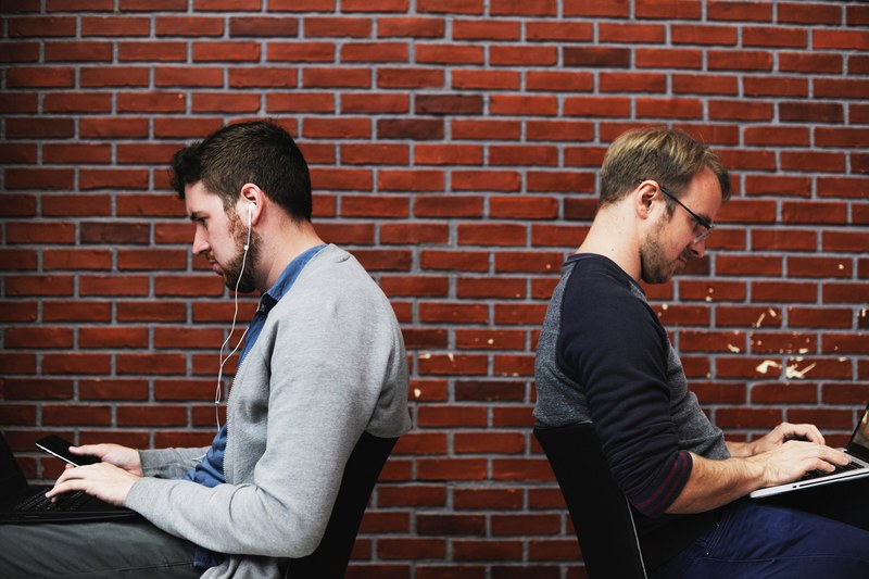two men working back to back outside with a brick wall background, working separately on laptops 4k