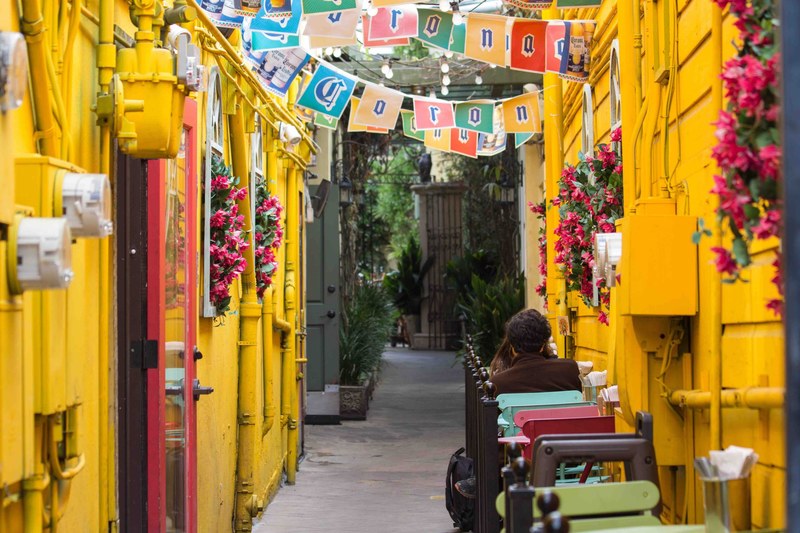 an alley between yellow walls and buildings and people sitting in a cafe under a festival decorations, yellow alleyway 4k