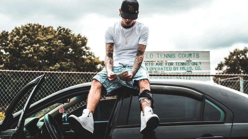 young man with edgy tattoos sitting atop his car couting stacks of money, counting his cash 4k