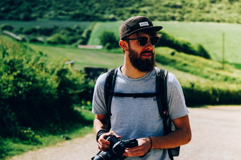 a bearded man wearing a backpack holds a camera outdoors in the isle of wight, lost on the white 4k